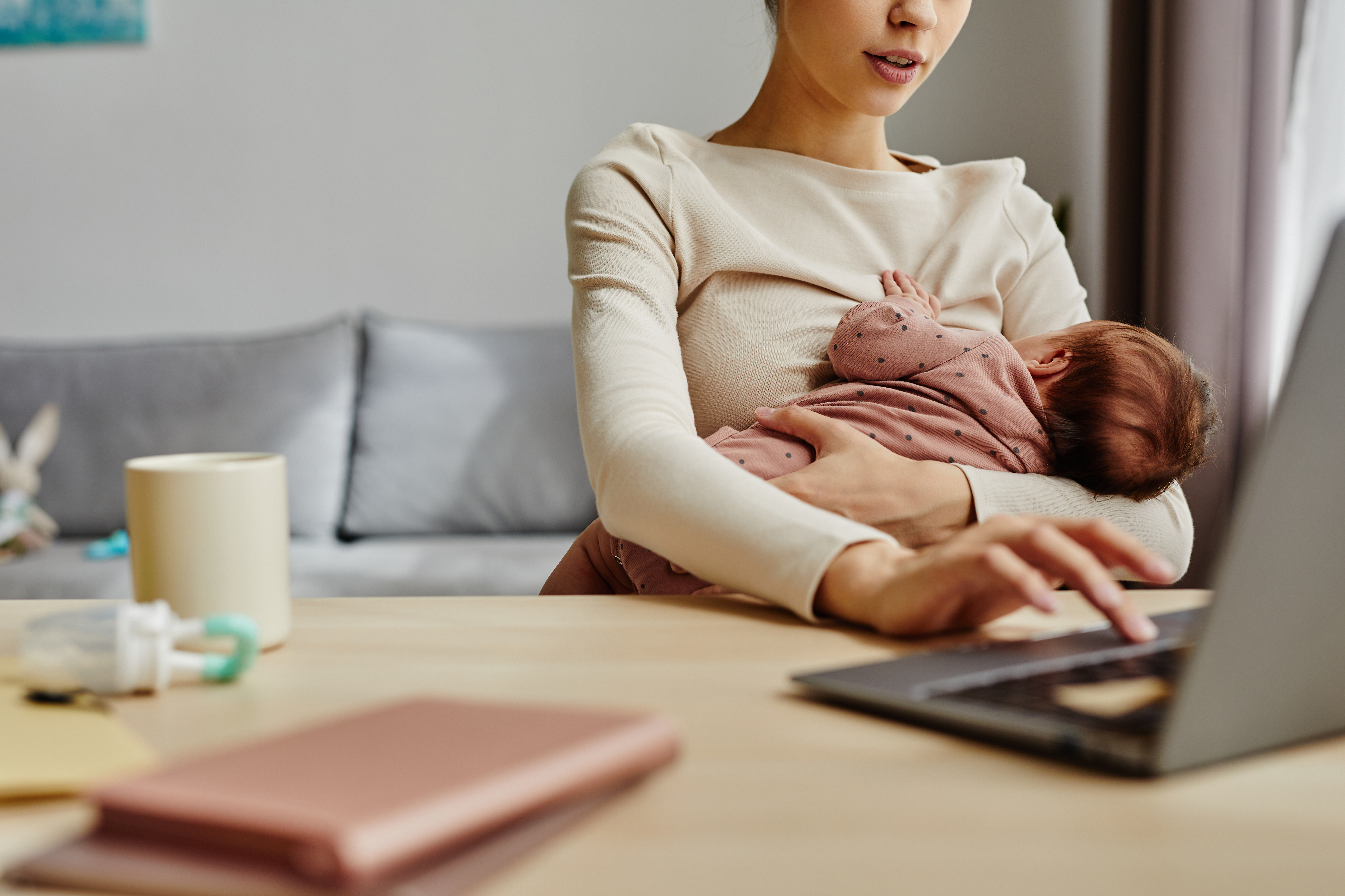 Cclose up of busy mother holding her baby while sitting at table and working on laptop at home